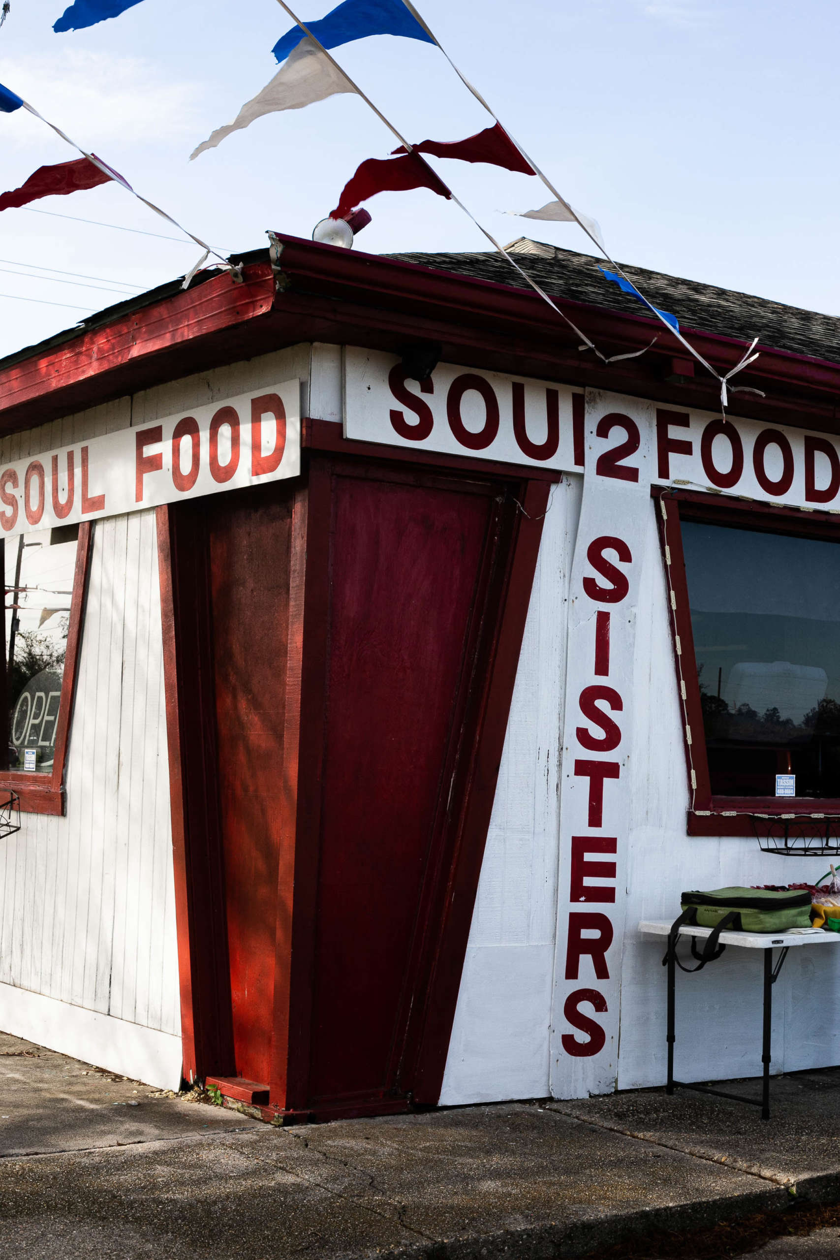 The corner of a red and white wooden building exterior. Intersecting panels along the roof and side read "2 Sisters Soul Food". Red, white, and blue triangular flags fly from streamers attached to the roof. A white plastic folding table sits in front of one window, with a green bag on top of it. The other window frames an unlit "Open" sign.