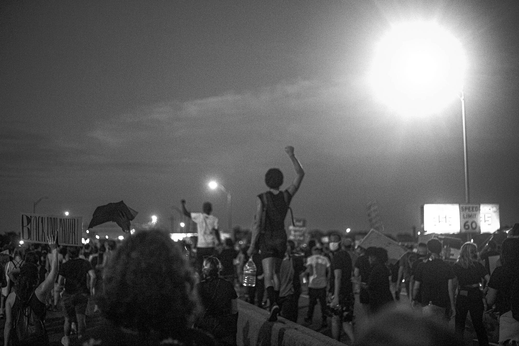 A black and white photo. Protesters walk together on a highway, taking up the width of the road; there are no cars to be seen. One protester, walking on the lane divider, raises their fist in the air and carries a water jug in their other hand. Another protester does the same, further down the road. A third protester raises a sign that says "Black Lives Matter" in capital letters. Light blazes from a streetlight in the corner of the scene.