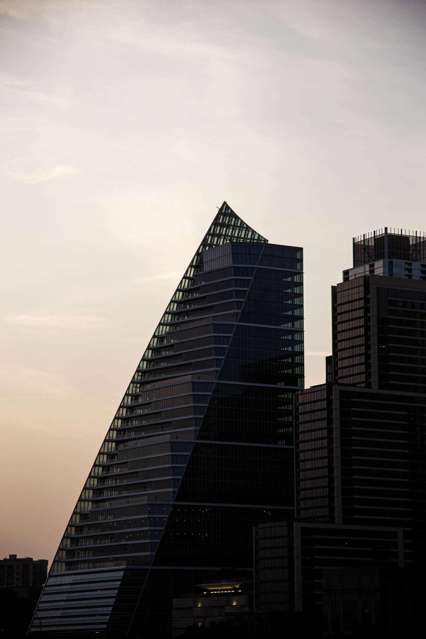 A tall building with a sloping triangular side stands in the dusk, almost silhouetted against a pale orange and blue sky with wispy clouds. More buildings stand behind it, and beneath it is a lighted entrance.