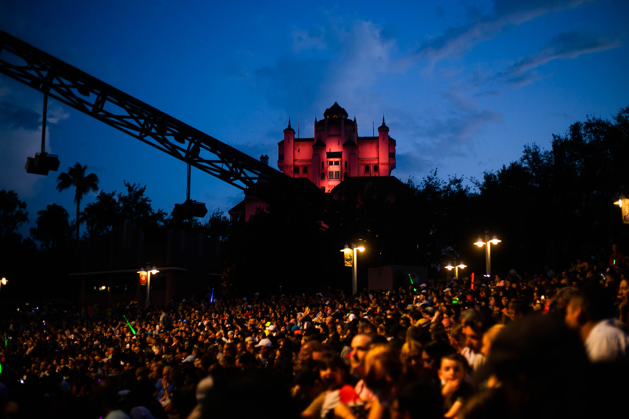 A sea of people at a concert, lit in gold by streetlights and by crane-mounted event lights above their heads. A treeline is silhouetted against the dark blue sky. On a hill above the treeline, a large building is lit in vivid red.