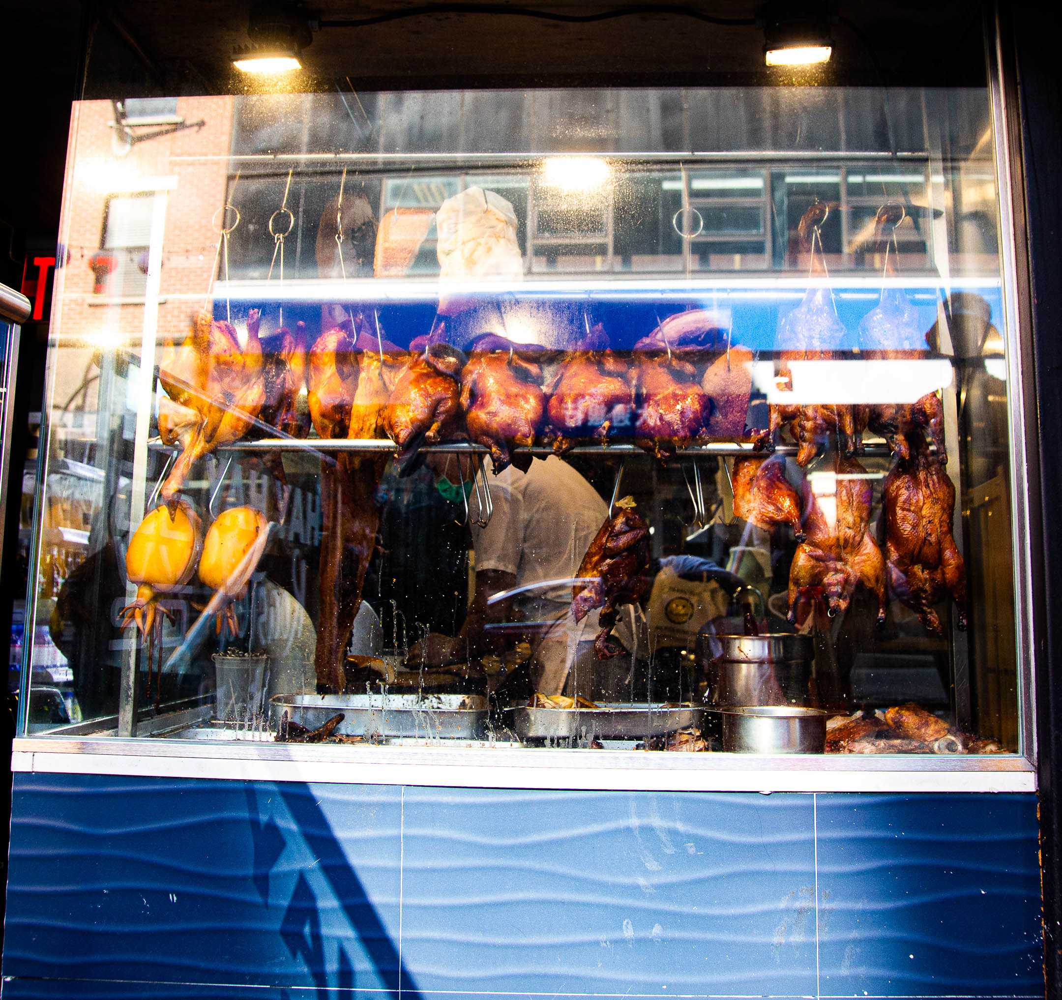 Various meats hang in a butcher's display case: duck, chicken, pork, and more. Lights from inside the butcher shop shine through the glass, and the apartment windows across the street reflect in the case. A person in a white shirt stands behind the case, working.