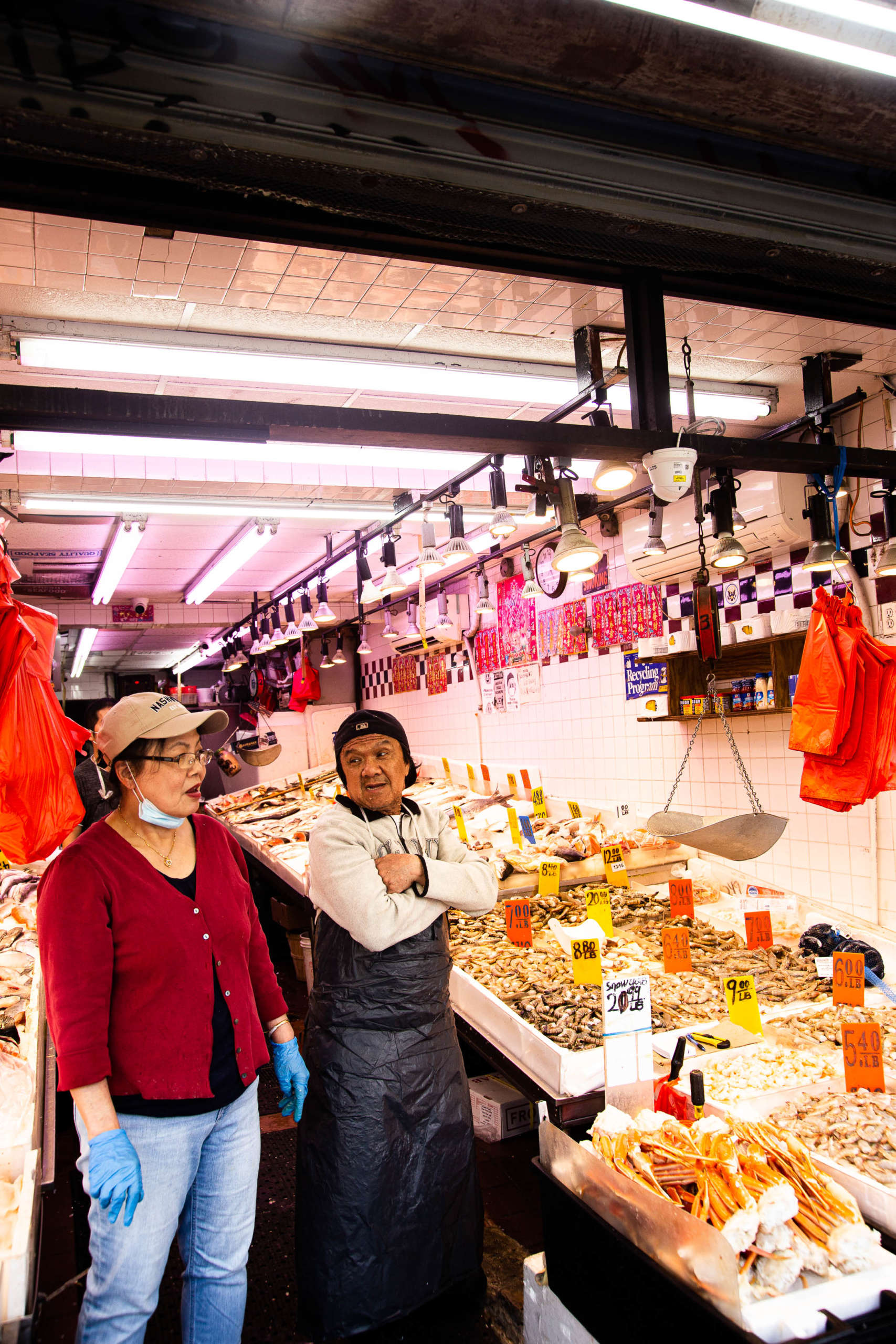 Two people standing in a seafood store, talking to each other. There are trays of seafood displayed all around them with orange and yellow price tags, and orange bags hanging above them.