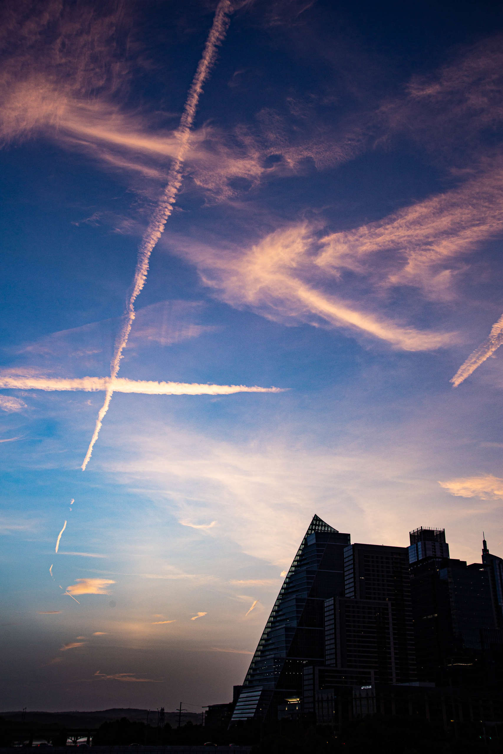 Wispy cirrus clouds, high in a blue sky, intersect with long white jet trails above the silhouette of a city skyline. The edges of the photo are darkened, with the clouds dimming to a vibrant pink.