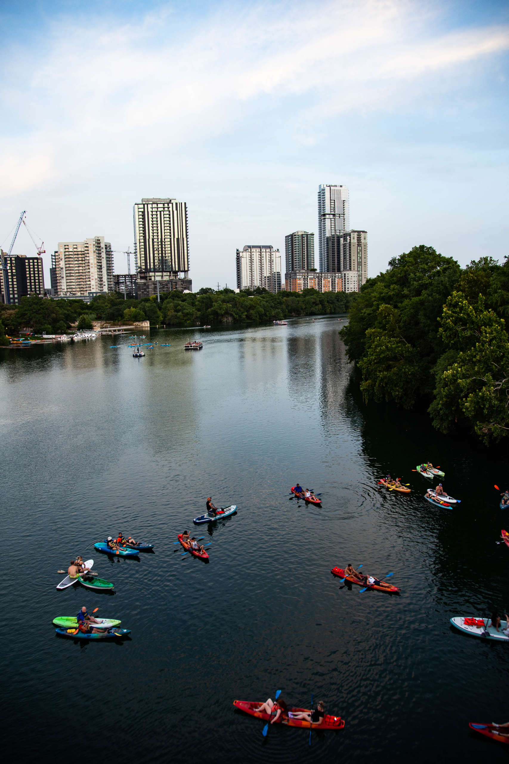 A group of people set out together in colorful kayaks on a river lined with trees. City buildings are visible in the background, reflecting on the water. The sky is bright blue with streaks of clouds.