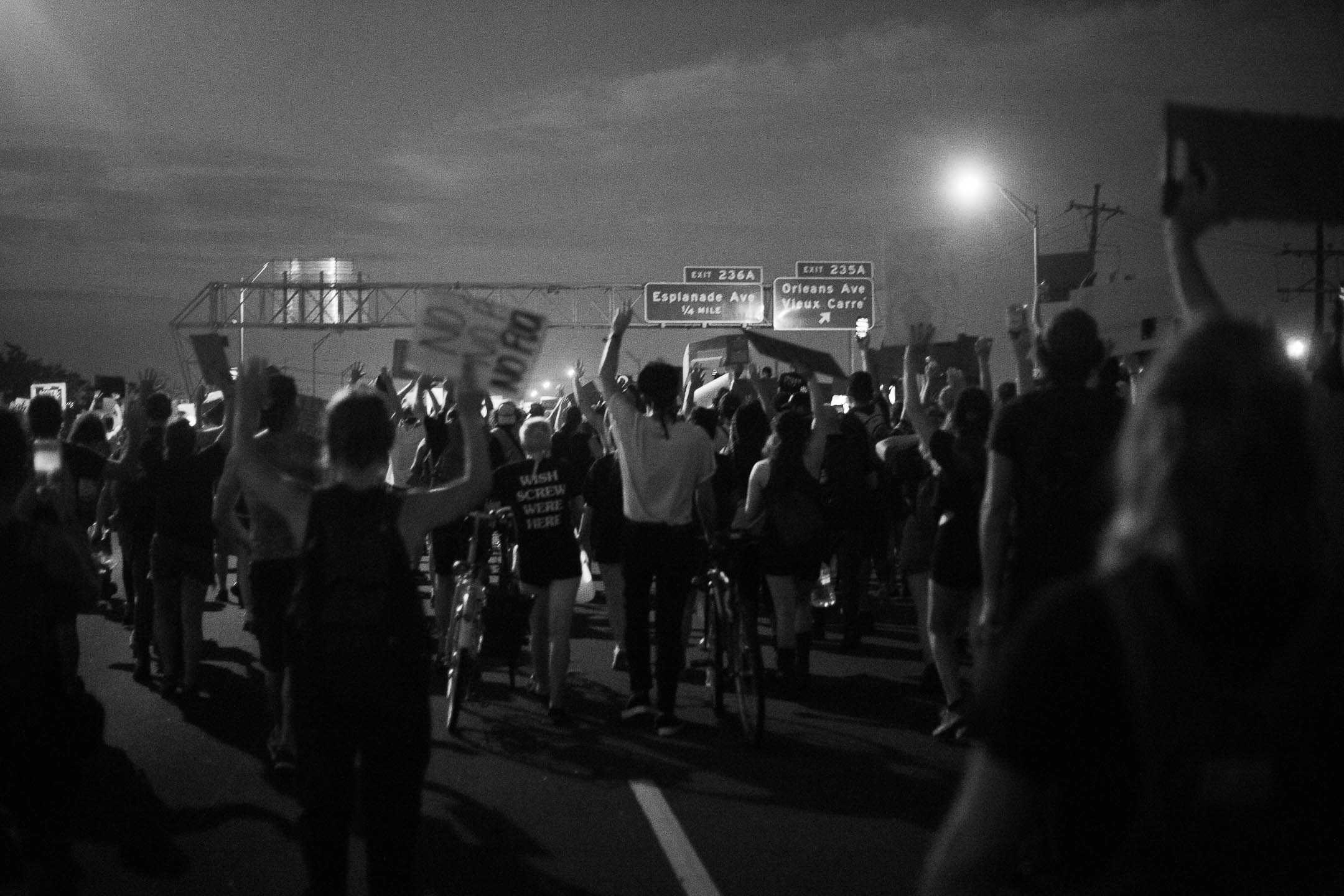 A black-and-white photo. A crowd of protesters fills a highway at night, lit by streetlights. They move together, on foot and on bikes, holding signs and raising fists. One person's shirt reads "Wish Screw Were Here". Above the protesters' heads, a highway gantry displays signs for Esplanade Ave and Orleans Ave / Vieux Carre.