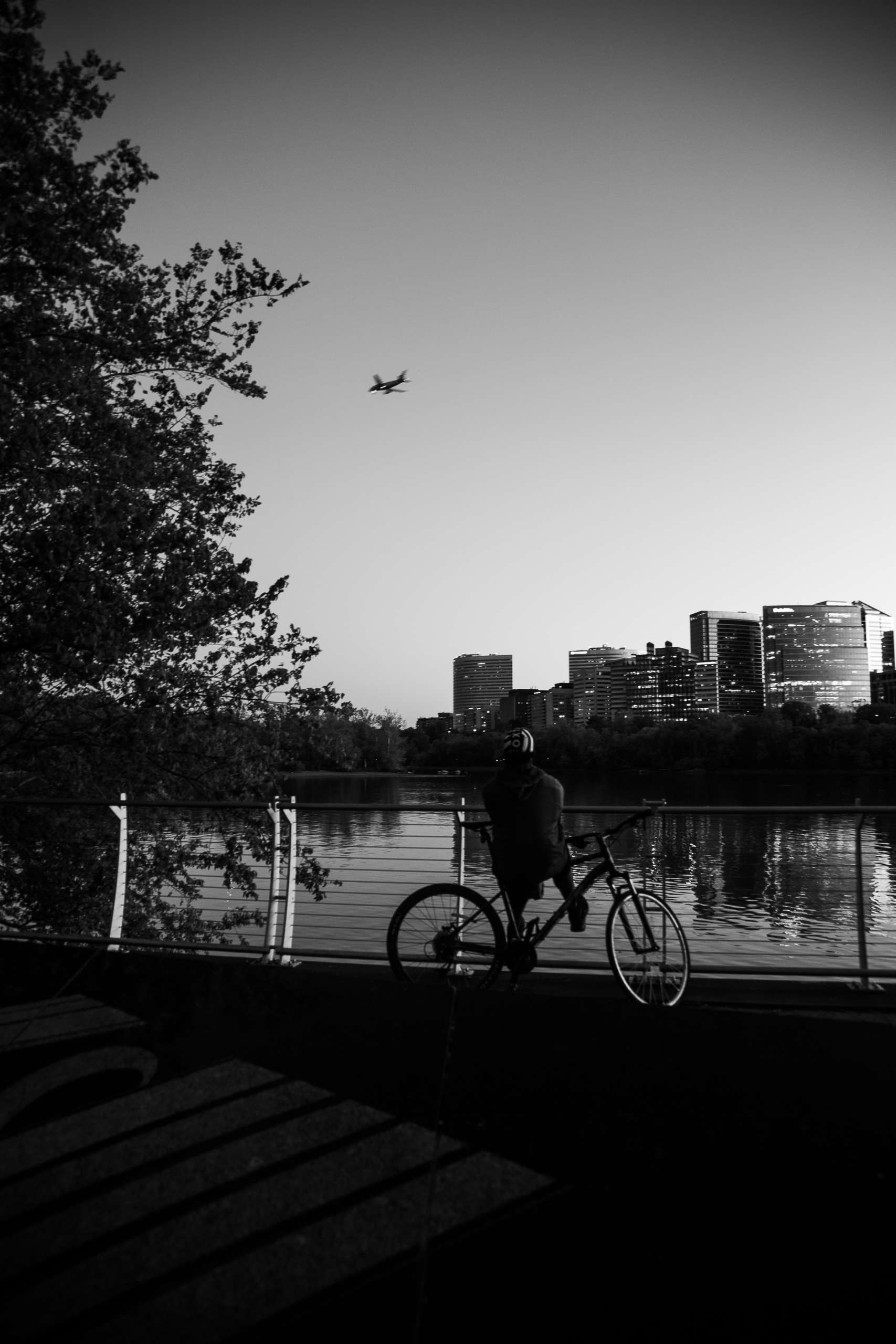 A black-and-white photo. A cyclist pauses by a guard rail overlooking a reflecting body of water, with downtown city buildings on the opposite bank. A plane flies low in the sky.