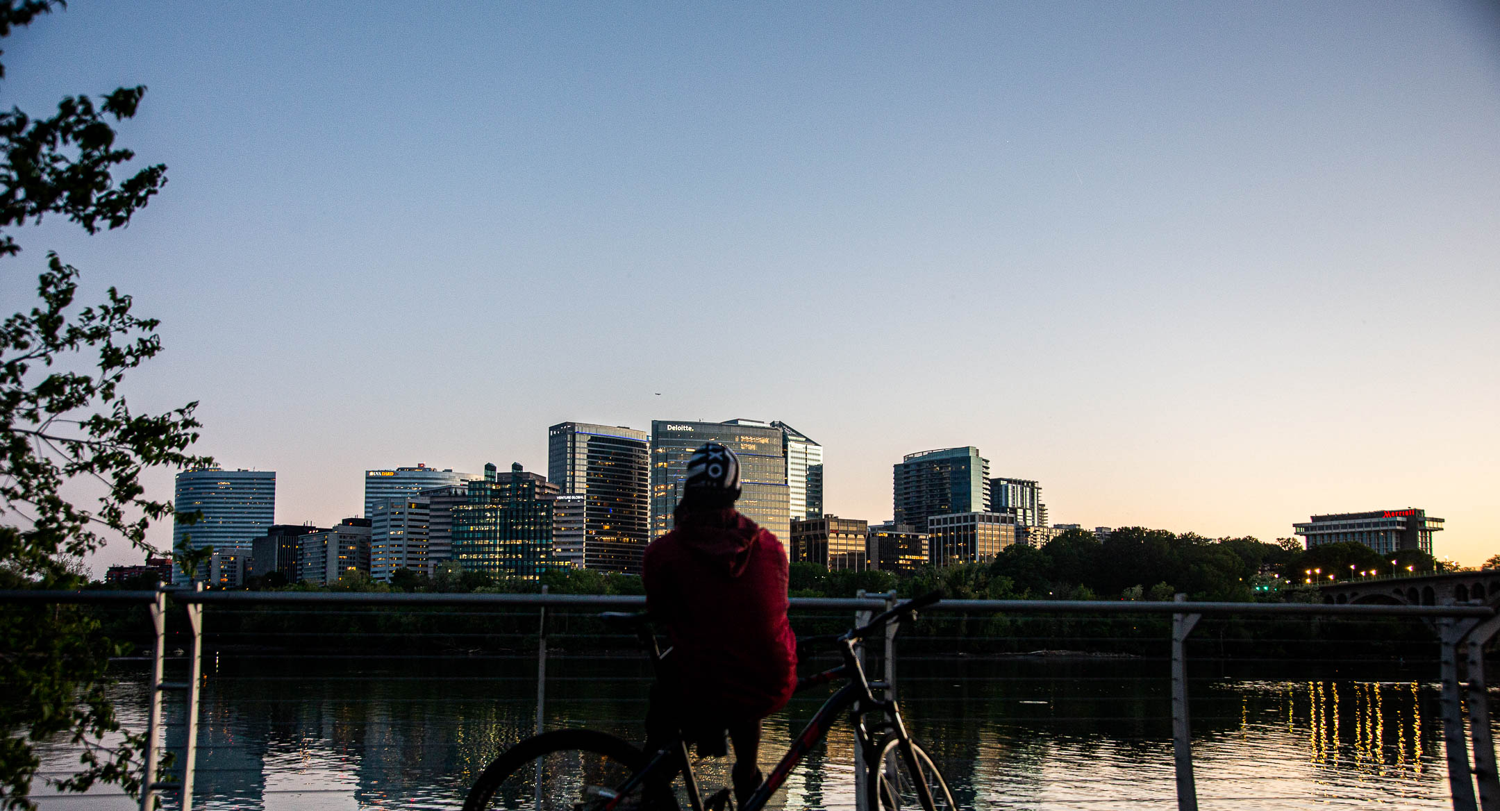 A cyclist in a red jacket looks out towards a city skyline, water separating them and reflecting the evening lights of the buildings.