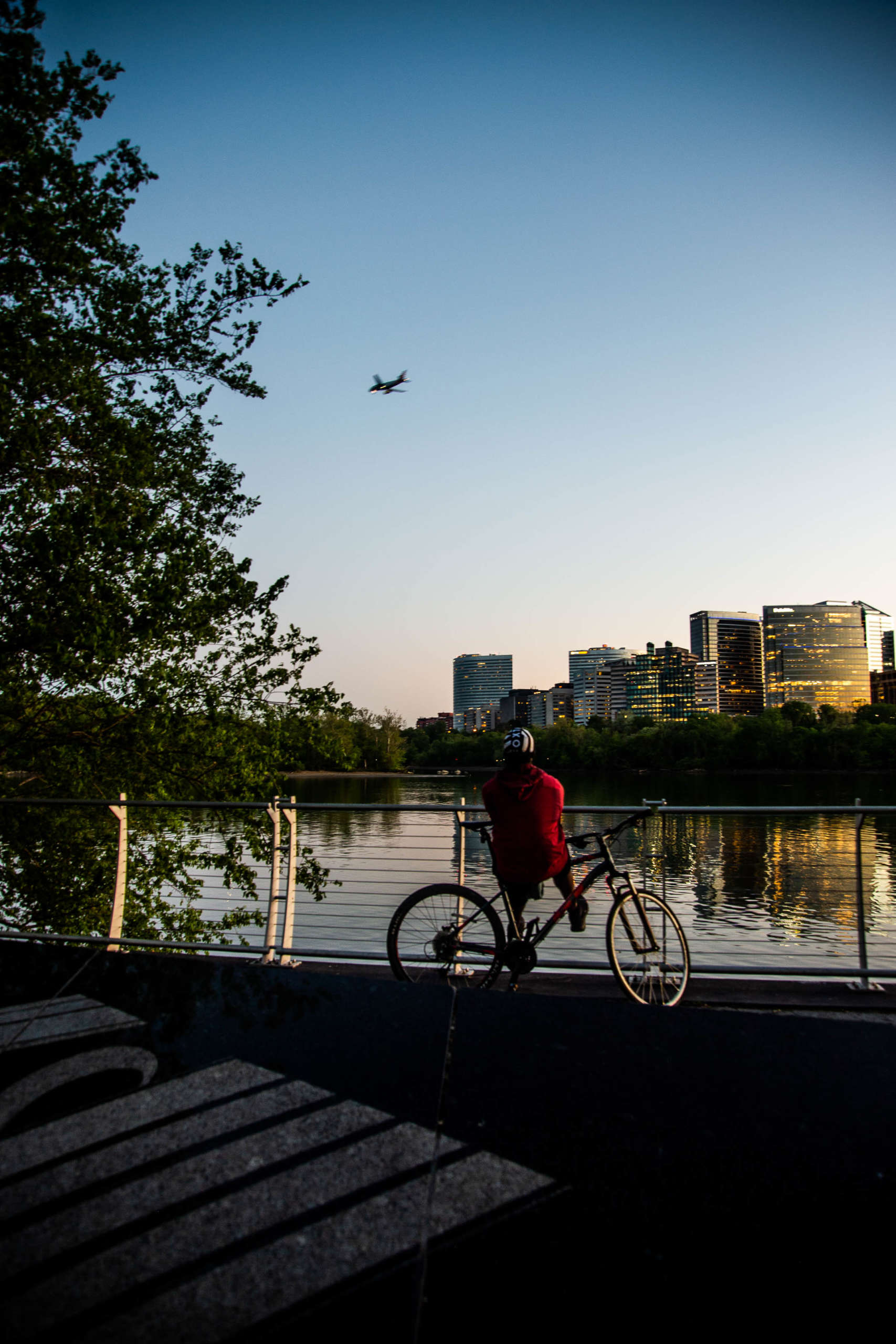 A cyclist pauses by a guard rail above a waterfront as they watch a plane soar overhead. Across the water is a city skyline, lit up in the gold of evening sunlight.