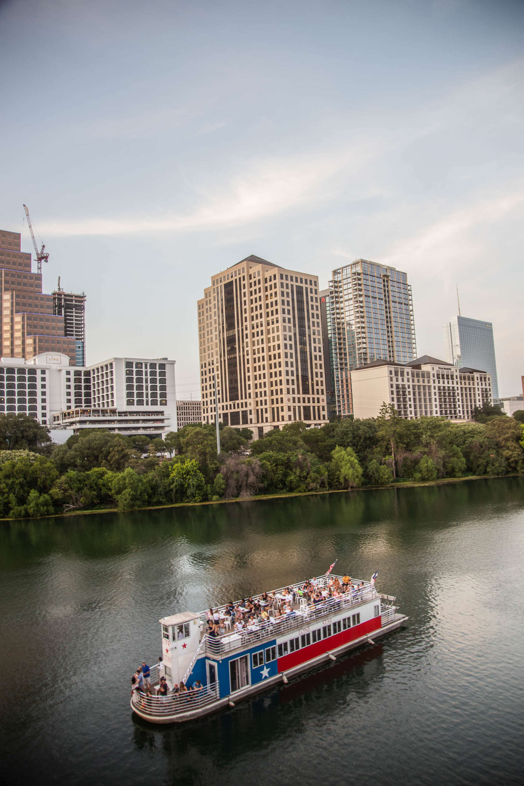 A ferryboat, decorated like the Texas flag, glides over a river. Its open top deck is packed with people, and several more stand at the curved front rail. In the background, trees line the river, and city buildings stand tall against a soft blue sky streaked with white clouds.