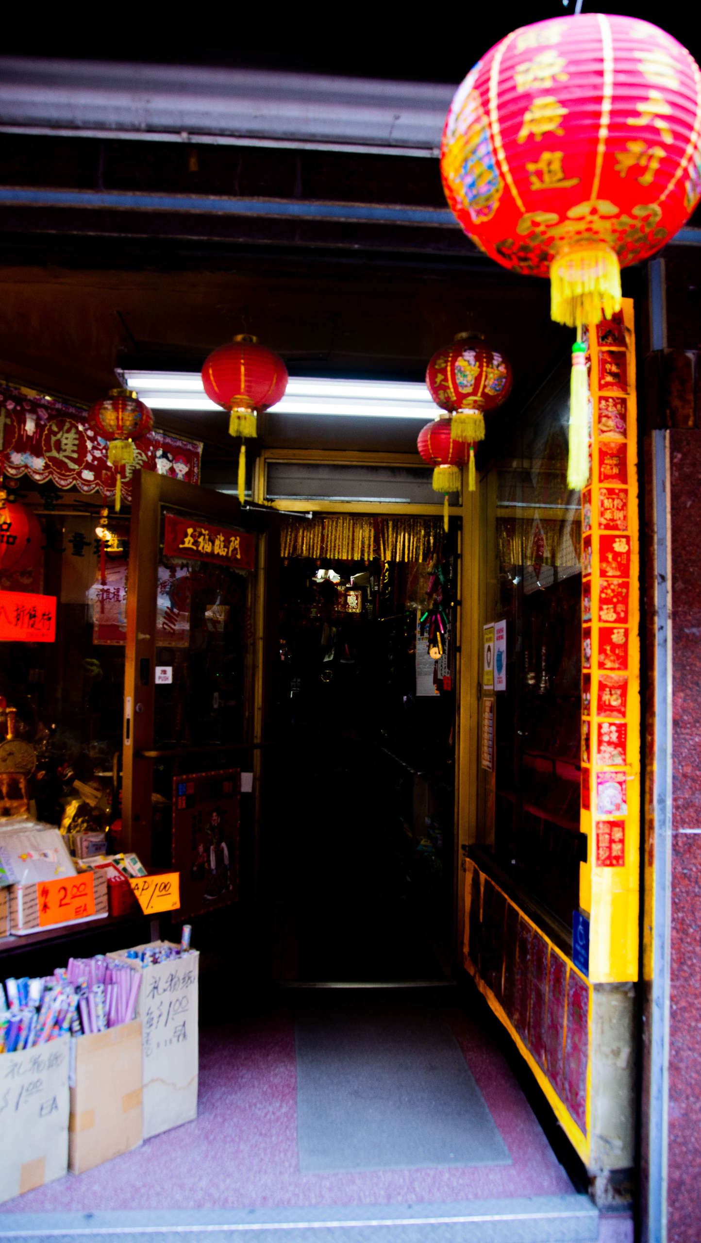 Red paper lanterns hang at the entrance to a storefront in Chinatown, NYC. Boxes outside the door are filled with merchandise and labeled with prices. The store is decorated in red and gold.