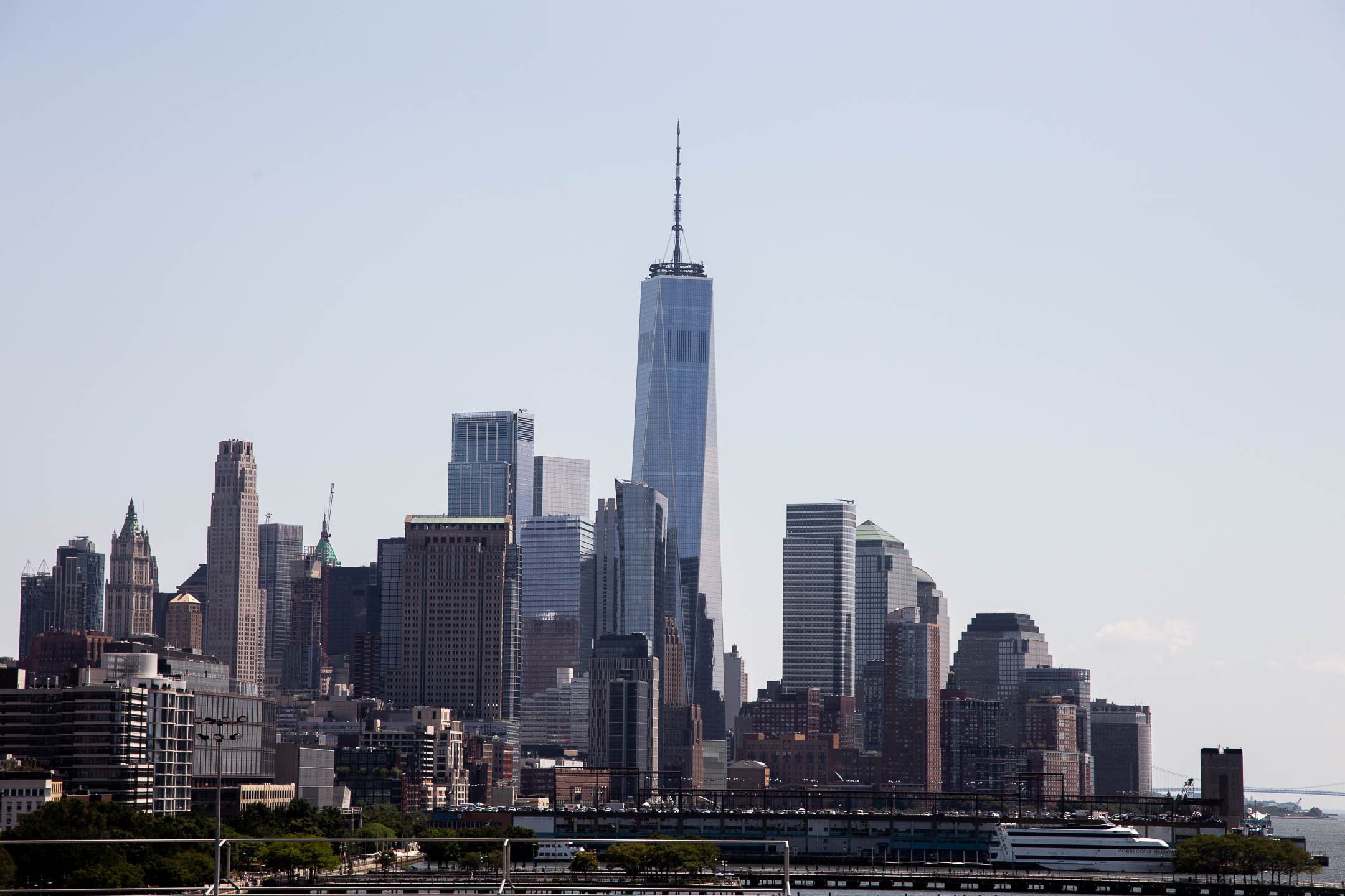 The New York City skyline, seen from a distance, with the soaring One World Trade Center building at the center. The skyscrapers reflect the soft blue sky and the evening sunlight. Down at the bottom of the photo, cars drive along a road above wharfs reaching out to the water.