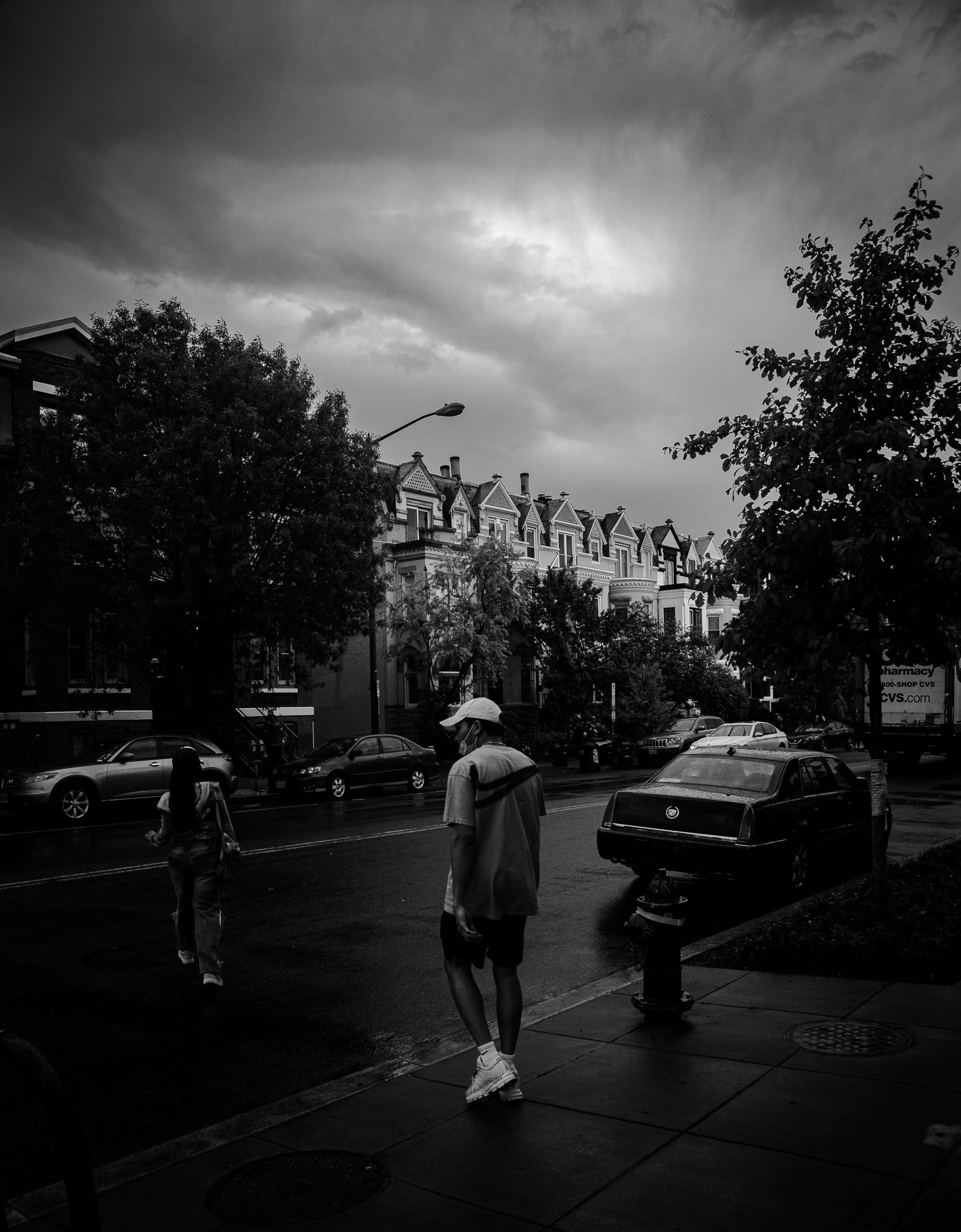 A black-and-white photo. A person in casual clothes and a baseball cap stands at the curb of a residential city street, waiting to cross. A second person with long hair has already started crossing the street. In the background are trees and a row of apartments with pointed roofs.