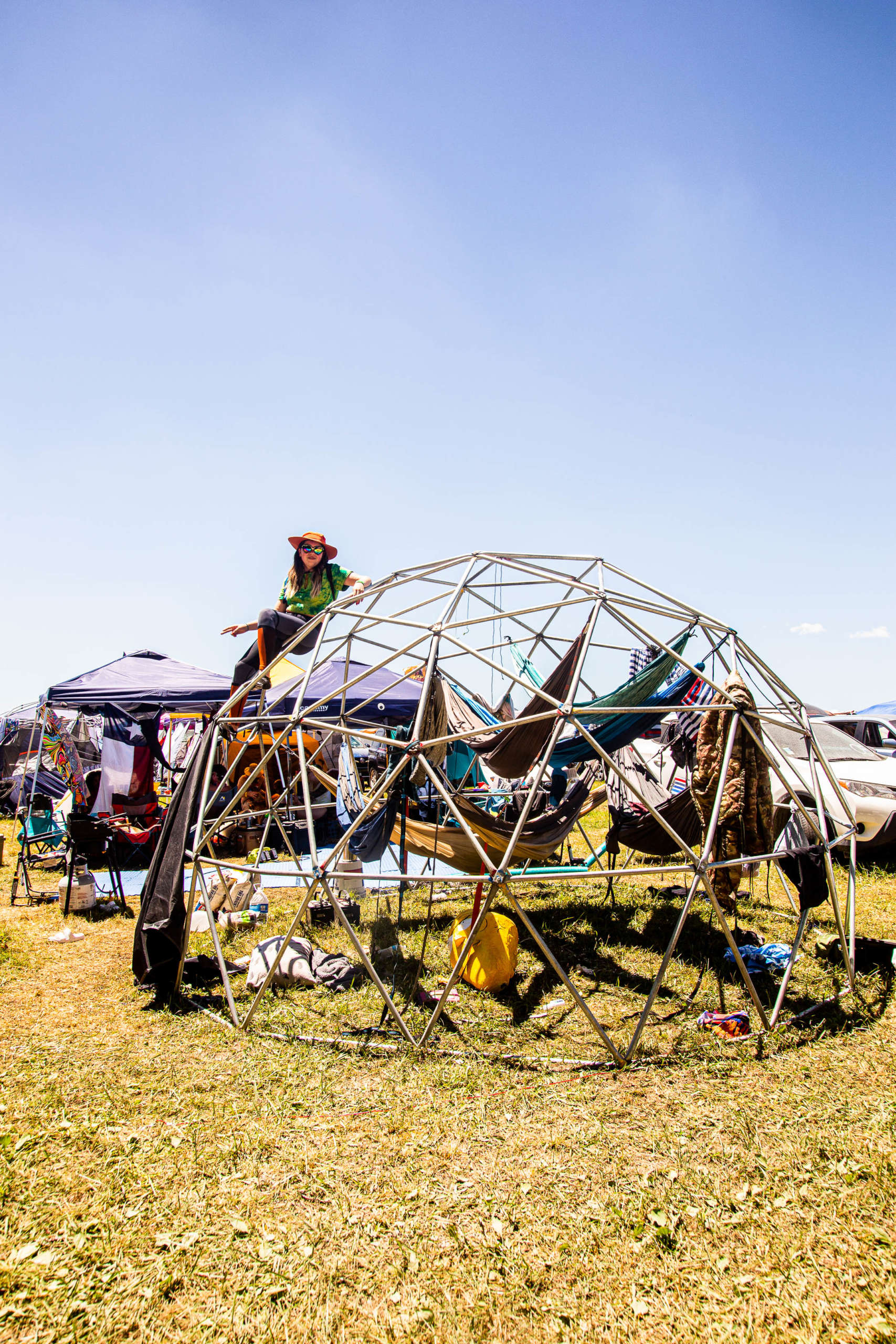 A person sits on top of a metal geodesic dome structure. Inside the dome are several multicolored hammocks hanging. Behind them are purple tents for an outdoor fair or event, with various products displayed for sale. It is a sunny summer day and cars are parked behind the tents.