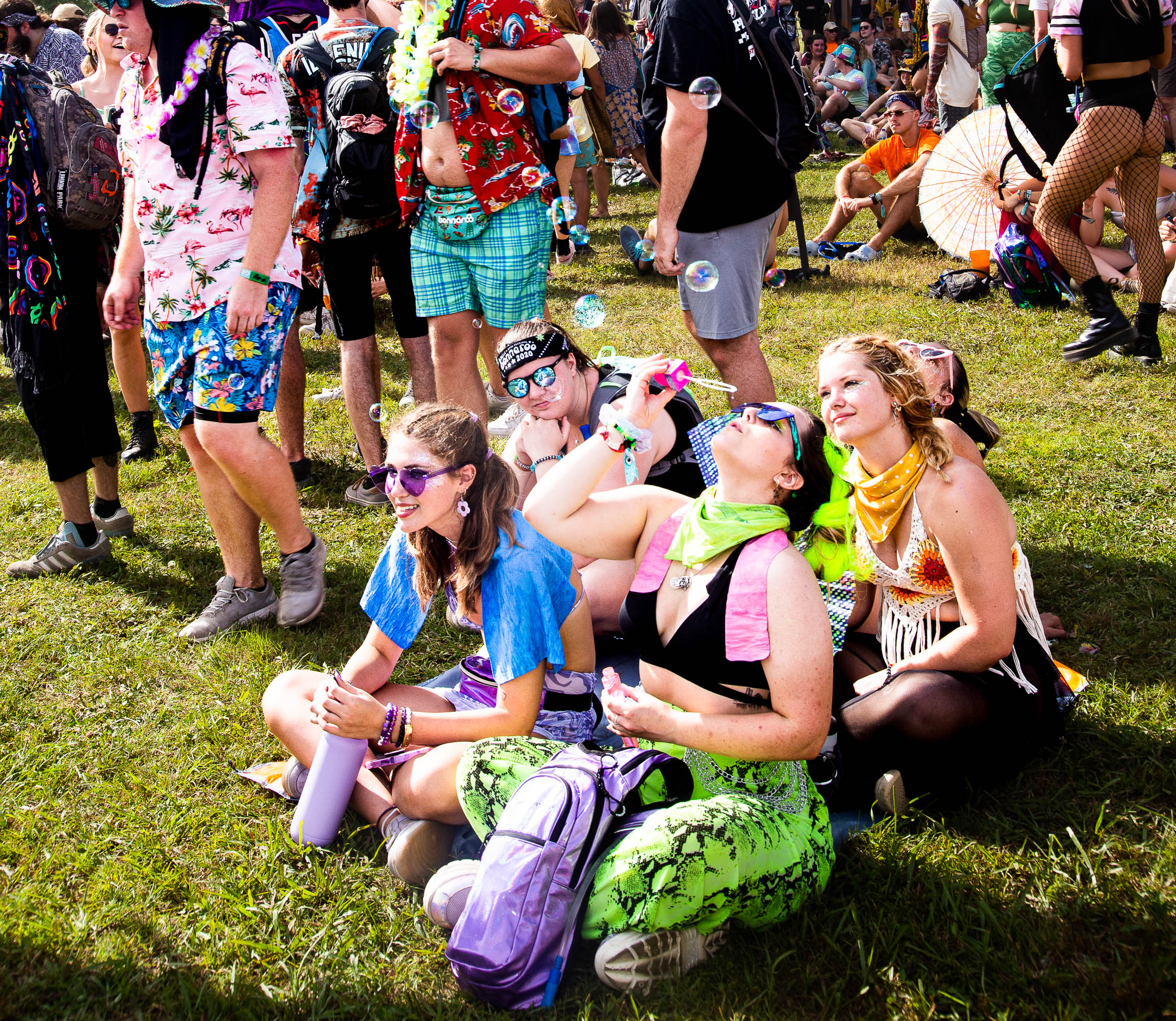 People waiting outdoors at an event, seated and standing, dressed in colorful summer clothes: Hawaiian shirts, crocheted tops, sunglasses, parasols. A person seated on the grass tilts their head back and blows soap bubbles with a pink wand.