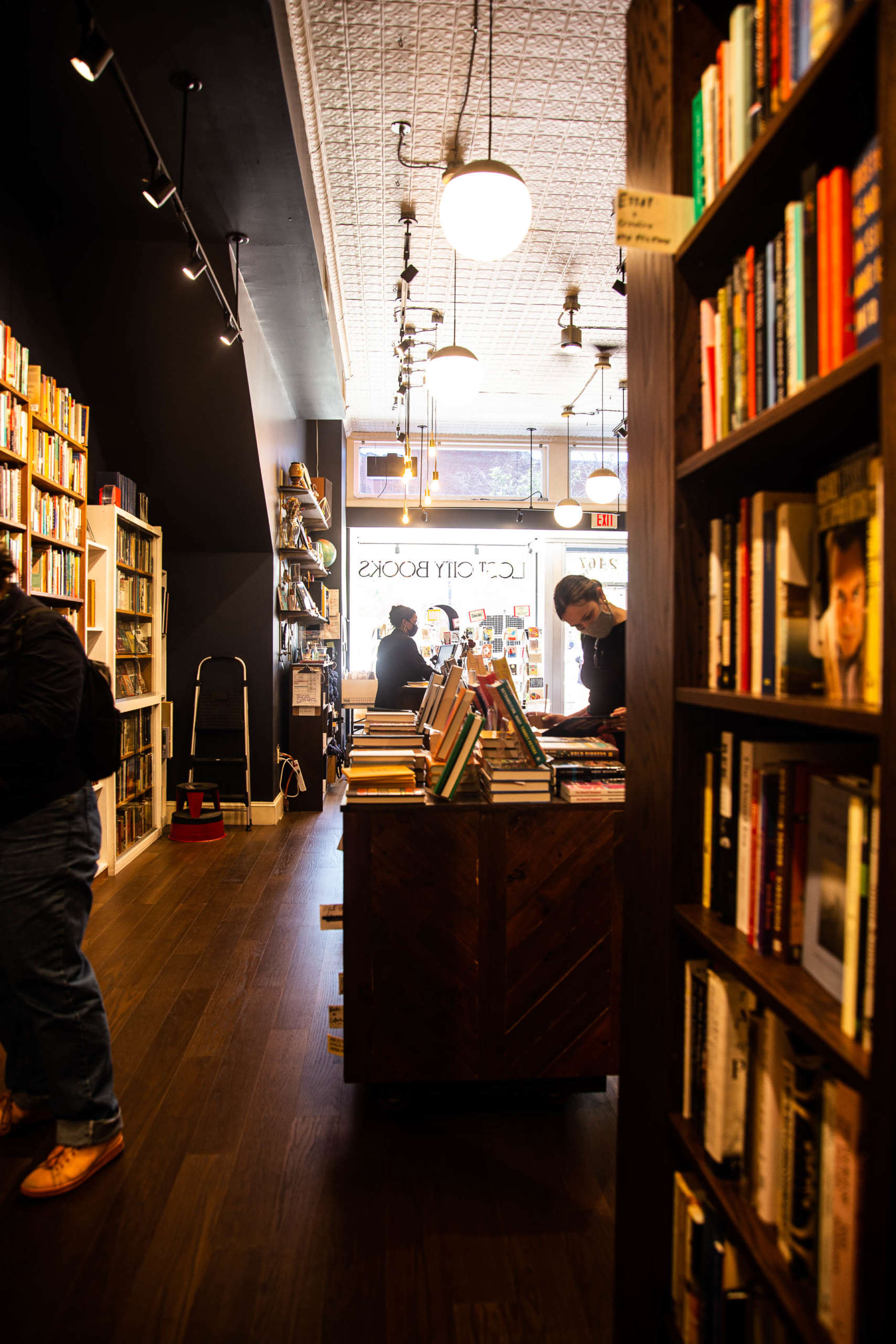 People in masks browse in a warmly-lit bookstore with wood floors and black walls. Reversed in the window is the store name "Lost City Books".