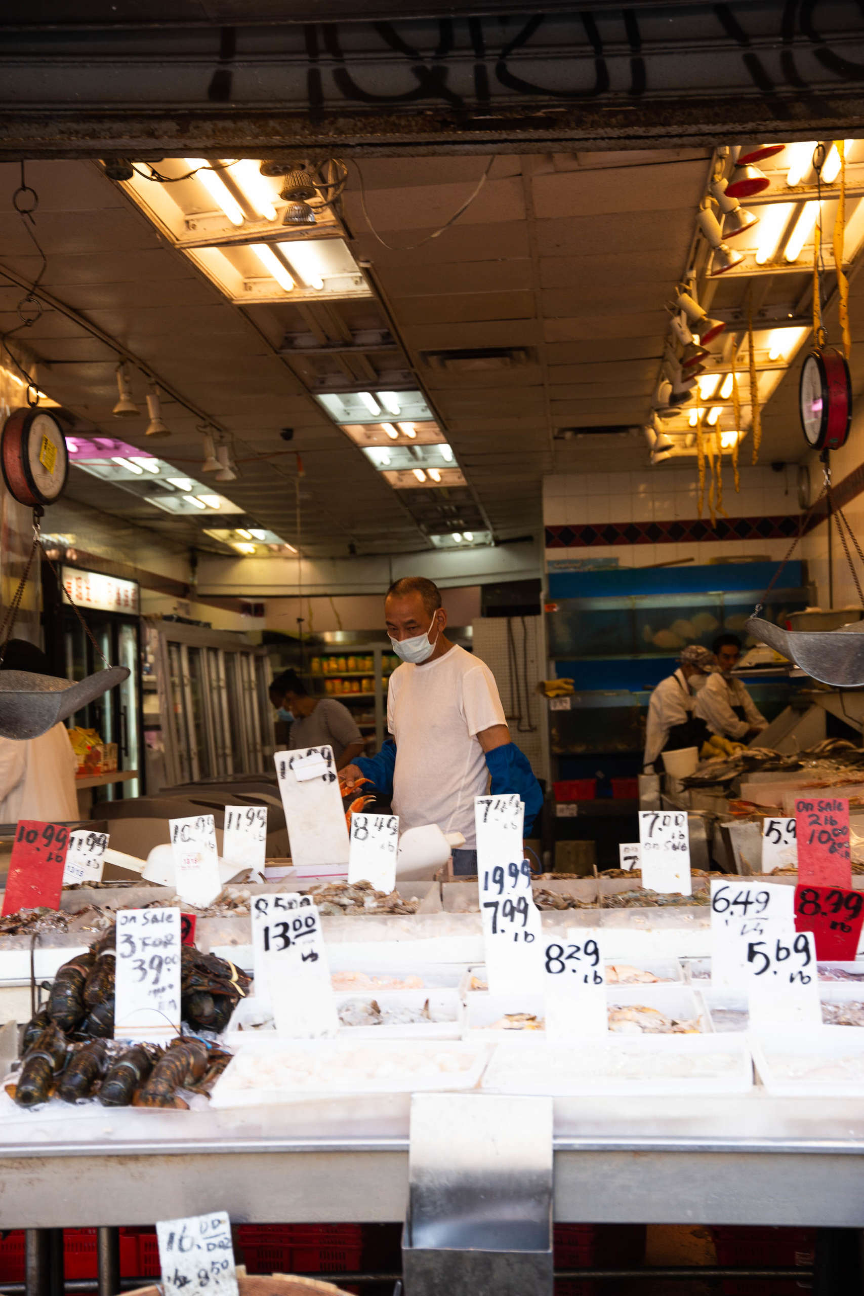 People browse the displays at a seafood store under fluorescent lighting. White price tags are arranged over each display.