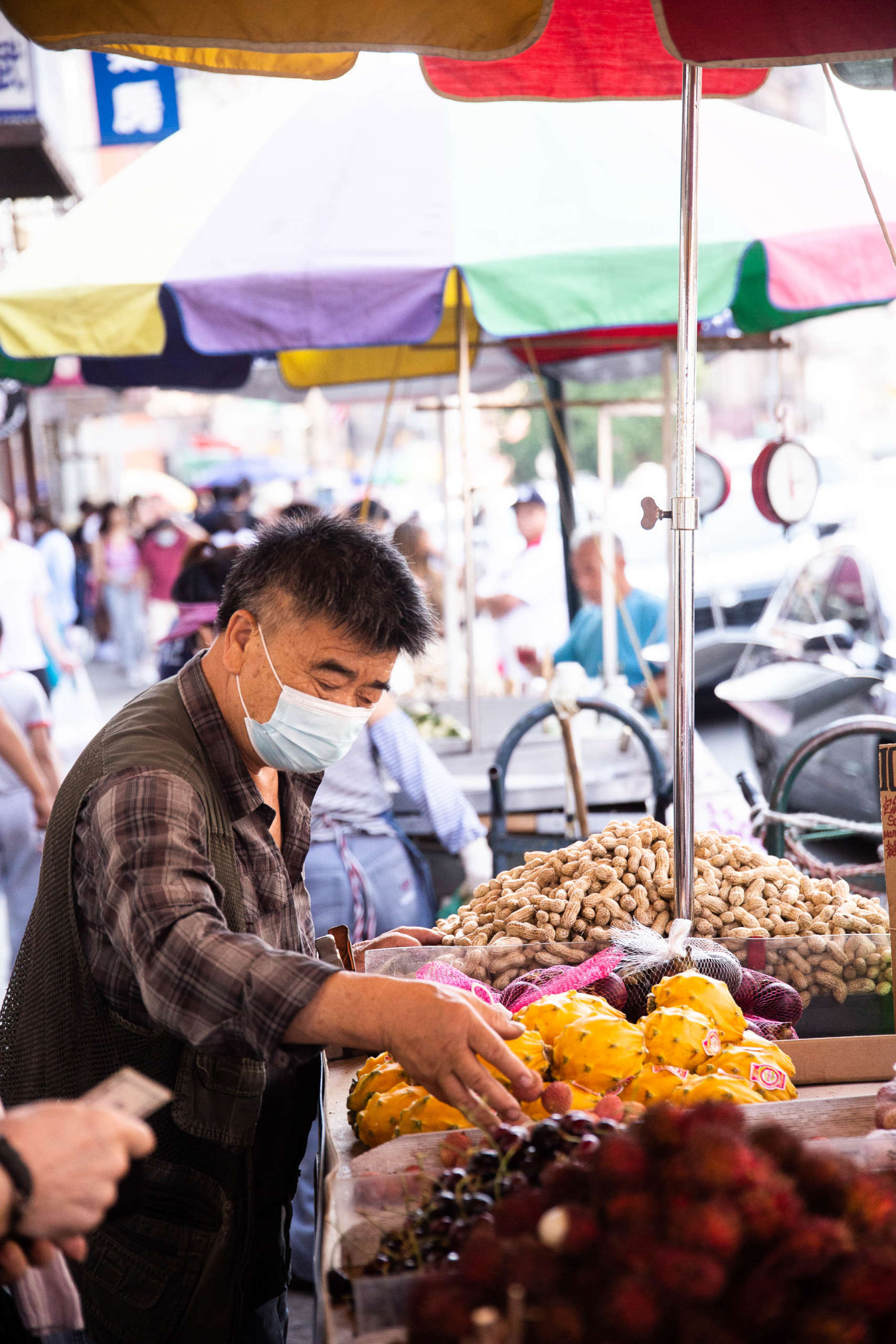 A person in a brown vest and shirt, wearing a blue surgical mask, thoughtfully selects a kiwano melon (or horned melon) from an outdoor food stand with a colorful umbrella above it. Other food is displayed nearby, including peanuts and cherries, and out-of-focus crowds move in the background.