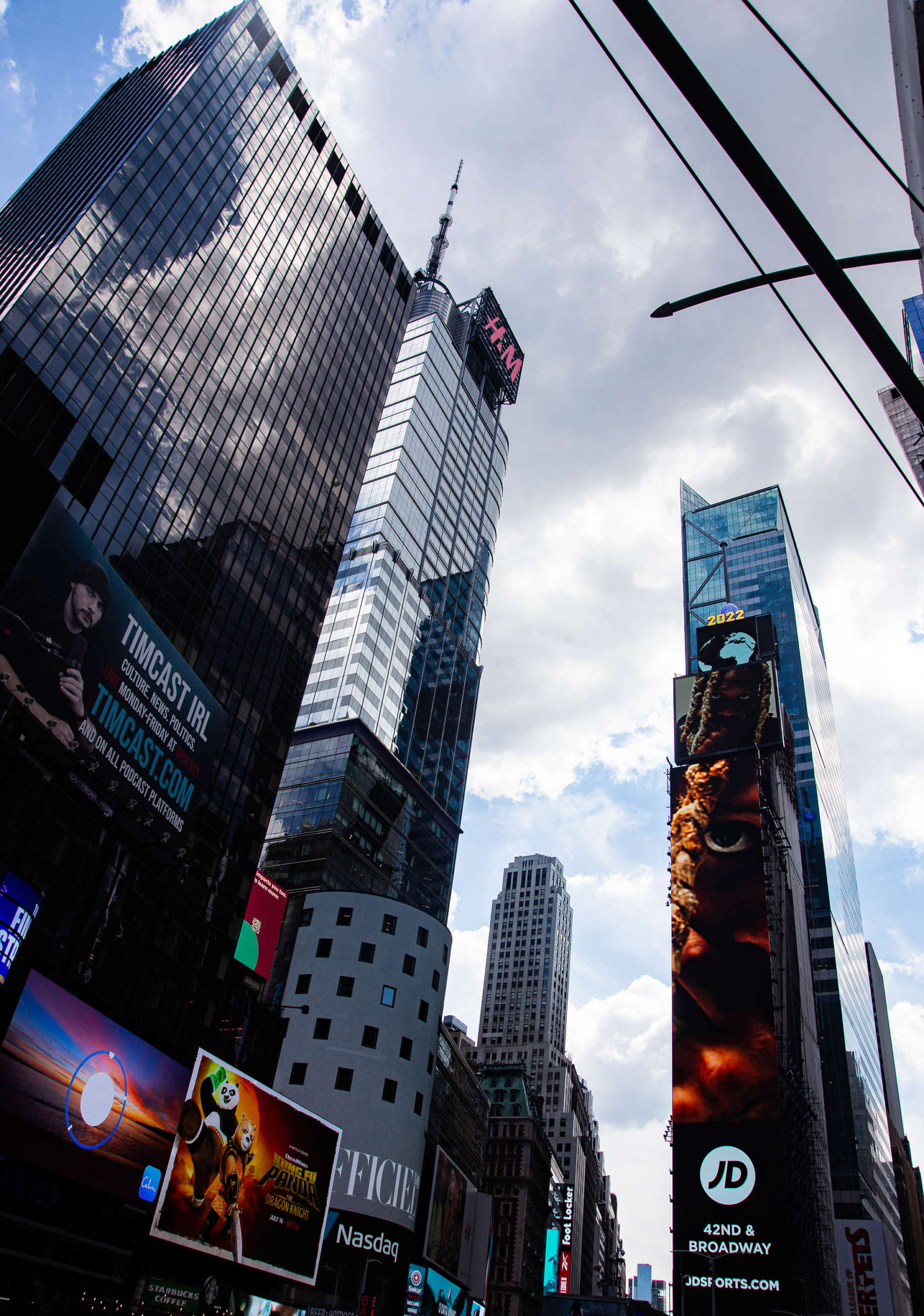 A low-angle shot of several tall buildings in Times Square, NYC. A large white cloud in the blue sky reflects off the mirrored windows of the buildings. The lower sections of the skyscrapers are covered in advertisements with various text: Timcast.com, H&M, Kung Fu Panda: The Dragon Knight, Nasdaq, 2022, JDSports, 42nd & Broadway. More billboards are visible on buildings as the line of the street vanishes into the distance.