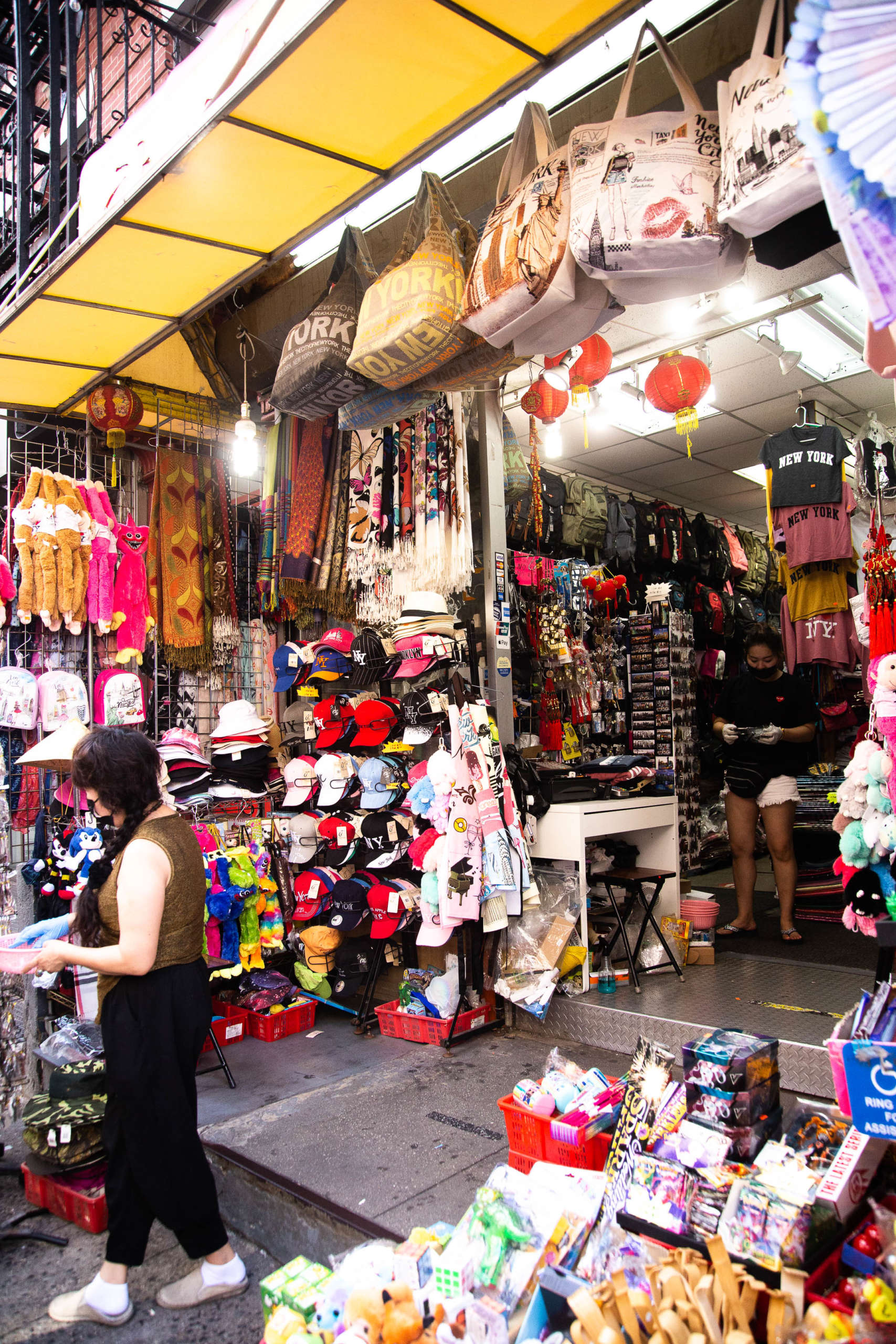 An open storefront in Chinatown, NYC, covered in unique gifts and souvenirs. Patterned tote bags hang from the roof next to paper lanterns; hats, scarves, puppets, toys, and red baskets of additional merchandise cover the walls and sit on tables. Inside, T-shirts, magnets, backpacks, and more are displayed. A person stands near the front of the open area, looking through a pink bowl; another stands inside the shop looking down at their phone.