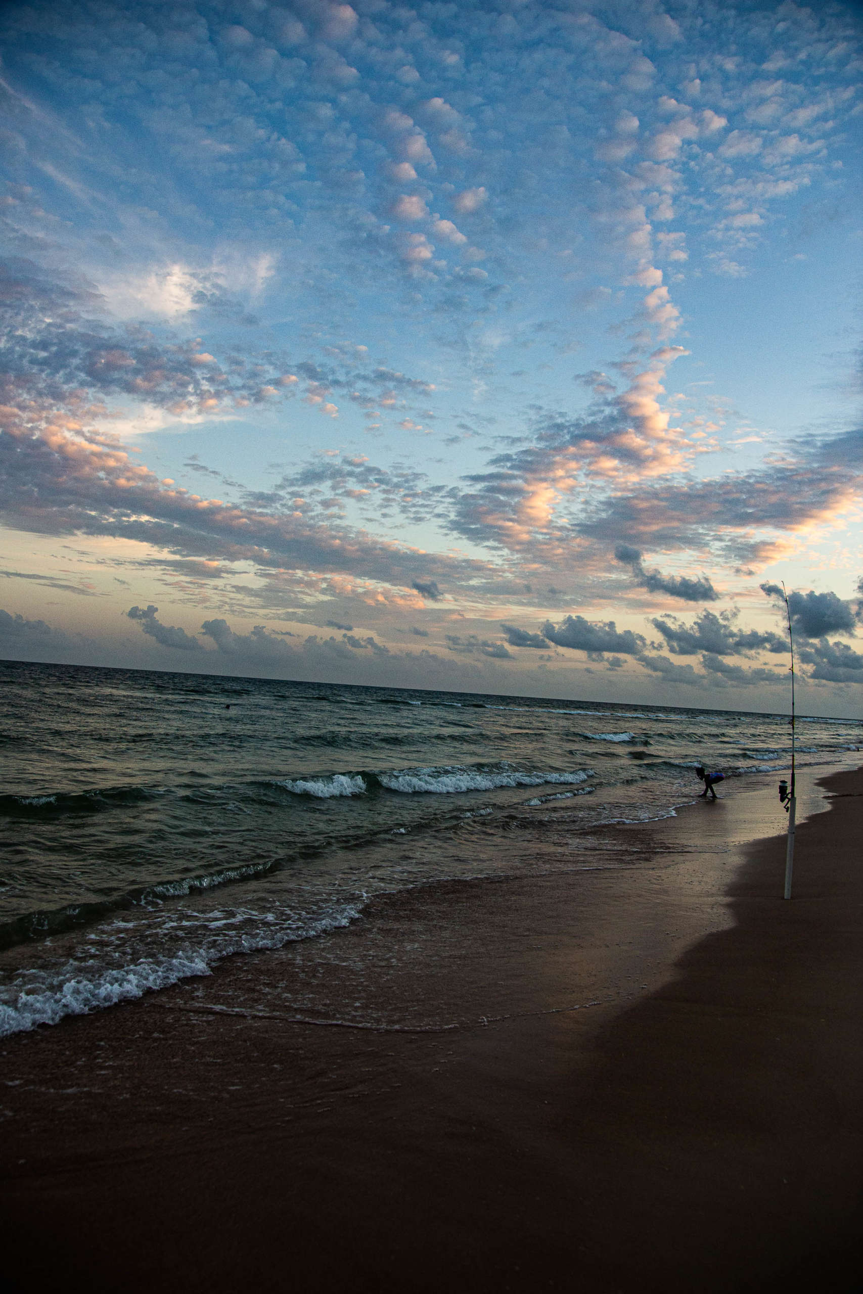 A beach just after sunset. Waves are crashing on the smooth shining sand, and a single person can be seen in the distance. Above the water are dozens of tiny scattered clouds, forming streaks and billows over the yellow-to-blue gradient of the sky.